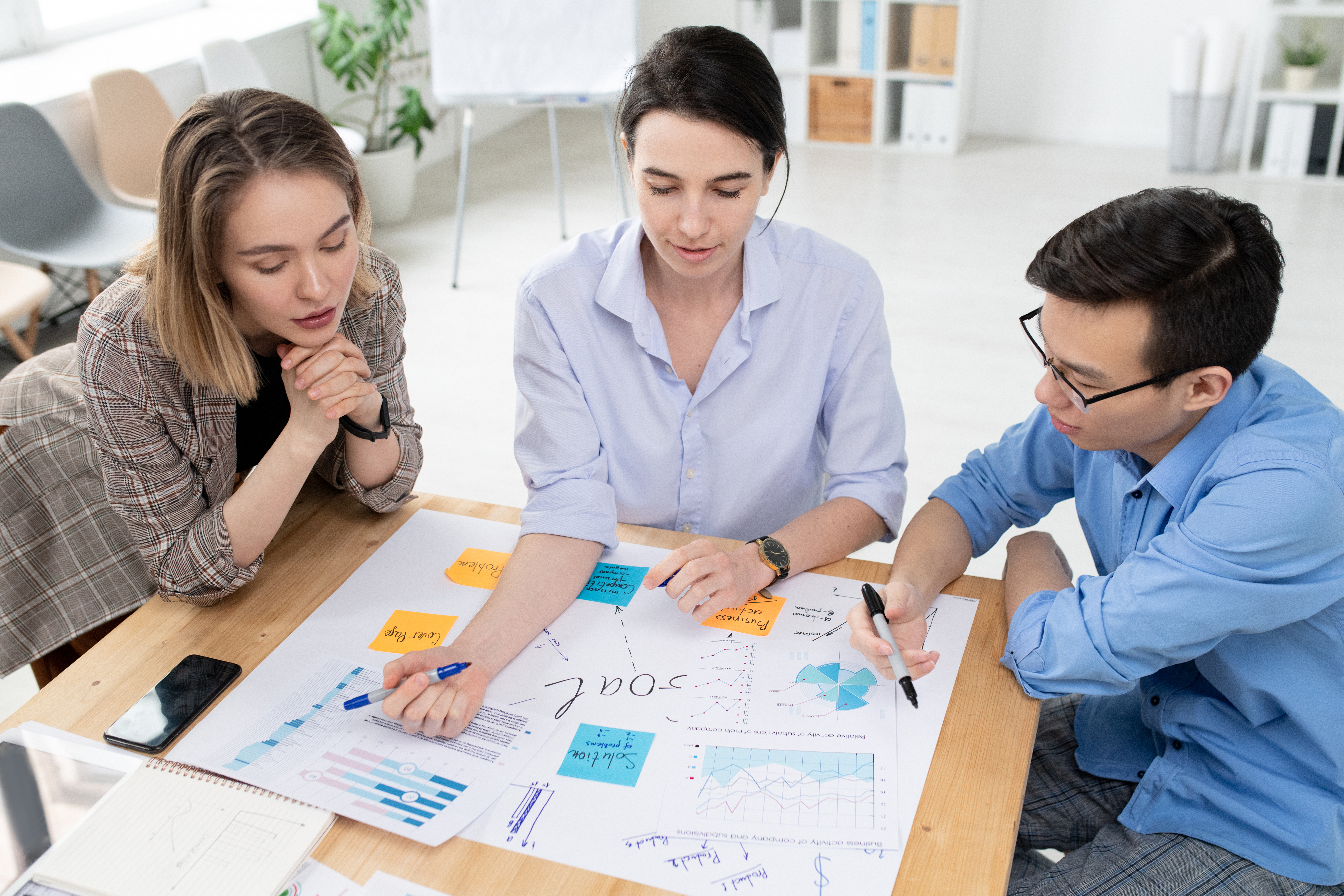 Two young women and businessman looking at large paper with business goals while brainstorming and discussing new ideas at meeting